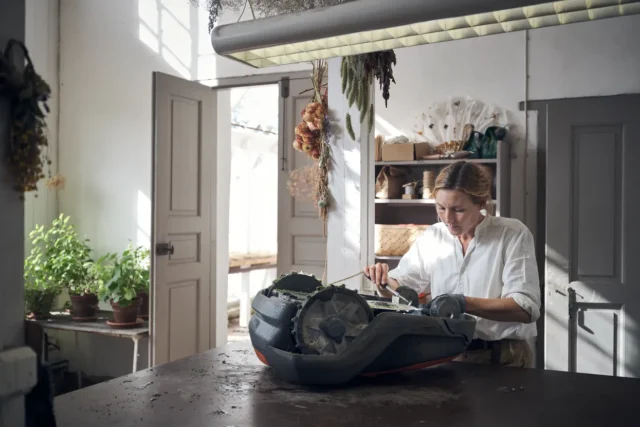 O persoană care lucrează la o mașină de tuns iarba robotizată într-un atelier, înconjurată de plante și unelte.