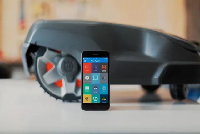 Un smartphone care afișează diverse aplicații lângă o mașină de tuns iarba robotizată.