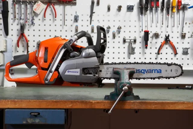 Vedere laterală a unui ferăstrău cu lanț Husqvarna fixat într-o menghină pe un banc de lucru, cu unelte pe un tablou perforat în spate.