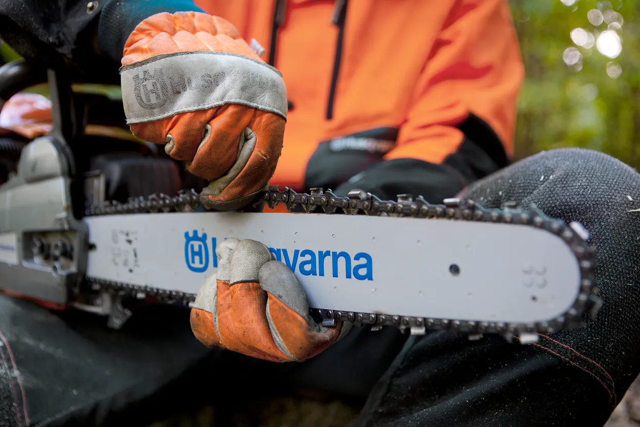 O persoană care poartă mănuși de lucru care reglează lanțul unui ferăstrău cu lanț Husqvarna.