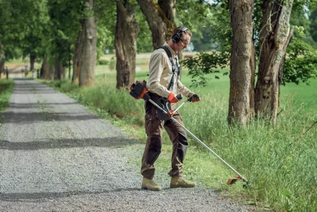 O persoană care folosește o mașină de tuns iarba pentru a tăia iarba de-a lungul unei cărări rurale mărginite de copaci.