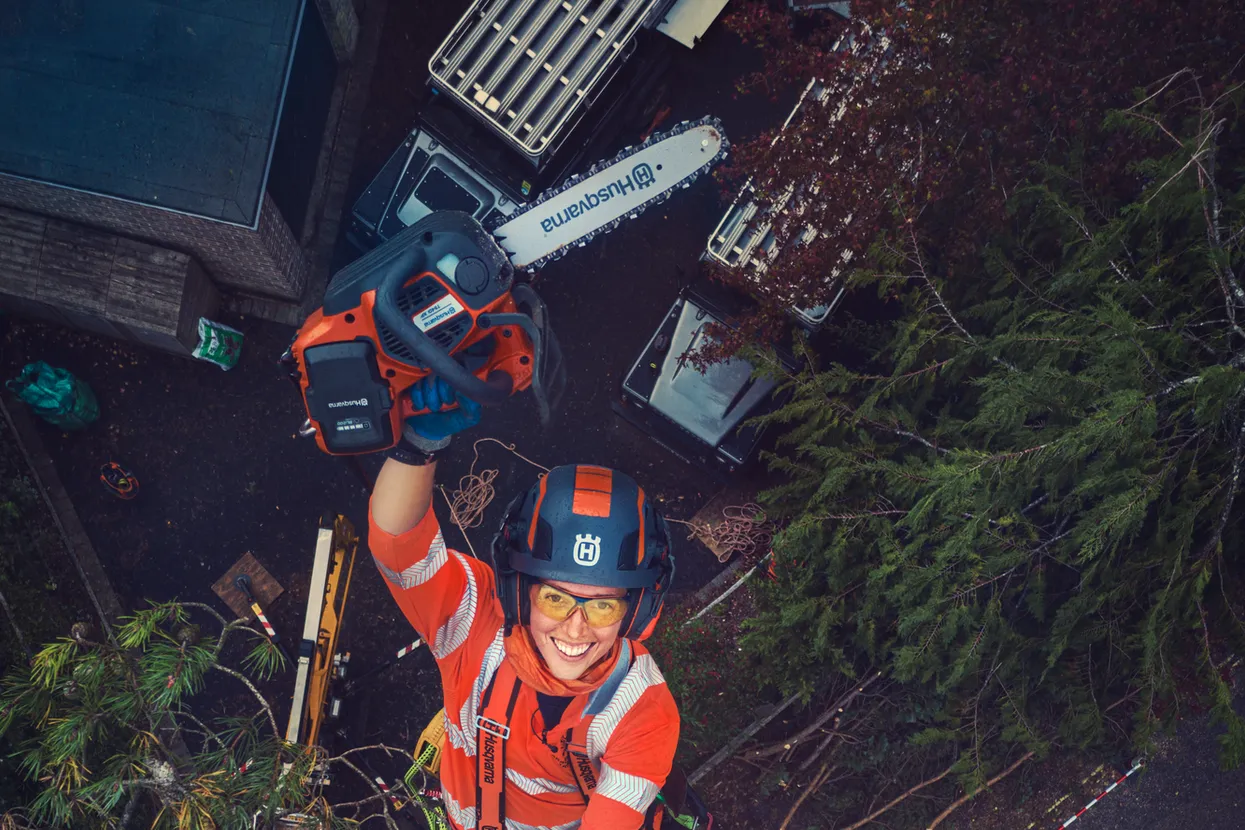 Arborist în echipament de lucru care se cațără într-un copac cu o drujbă și o cască.