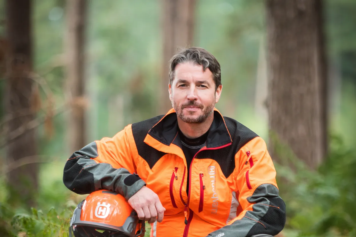 O persoană care stă pe un buștean de copac într-un cadru forestier, purtând echipament de lucru și ținând o cască.