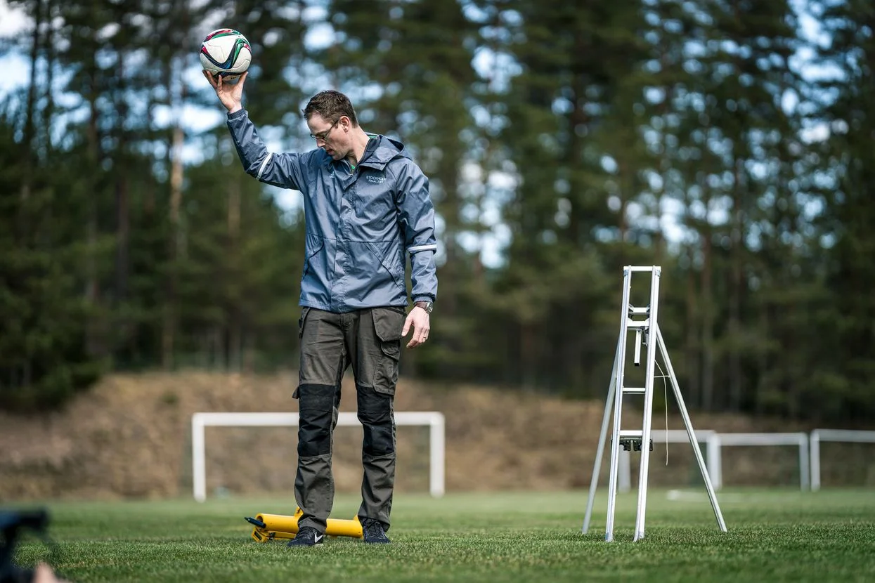 O persoană care stă pe un teren cu iarbă ținând o minge de fotbal, cu o scară în apropiere.