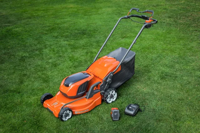 Mașină de tuns iarba cu baterii Husqvarna pe un gazon cu baterie și încărcător lângă ea.