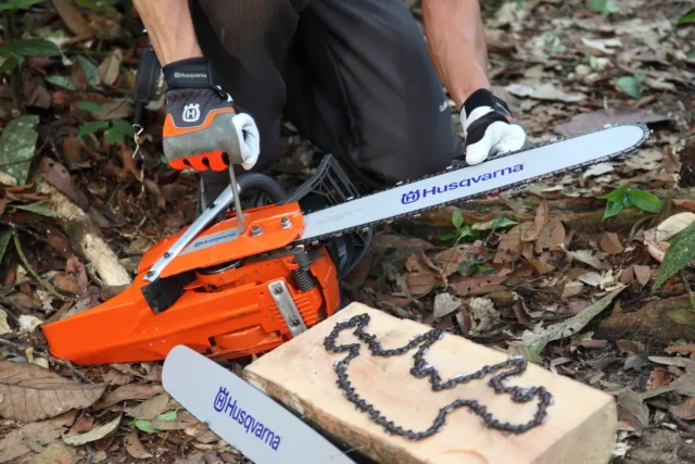 O persoană îngenuncheată într-o pădure, ajustând un ferăstrău cu lanț Husqvarna lângă un bloc de lemn cu un lanț de drujbă.