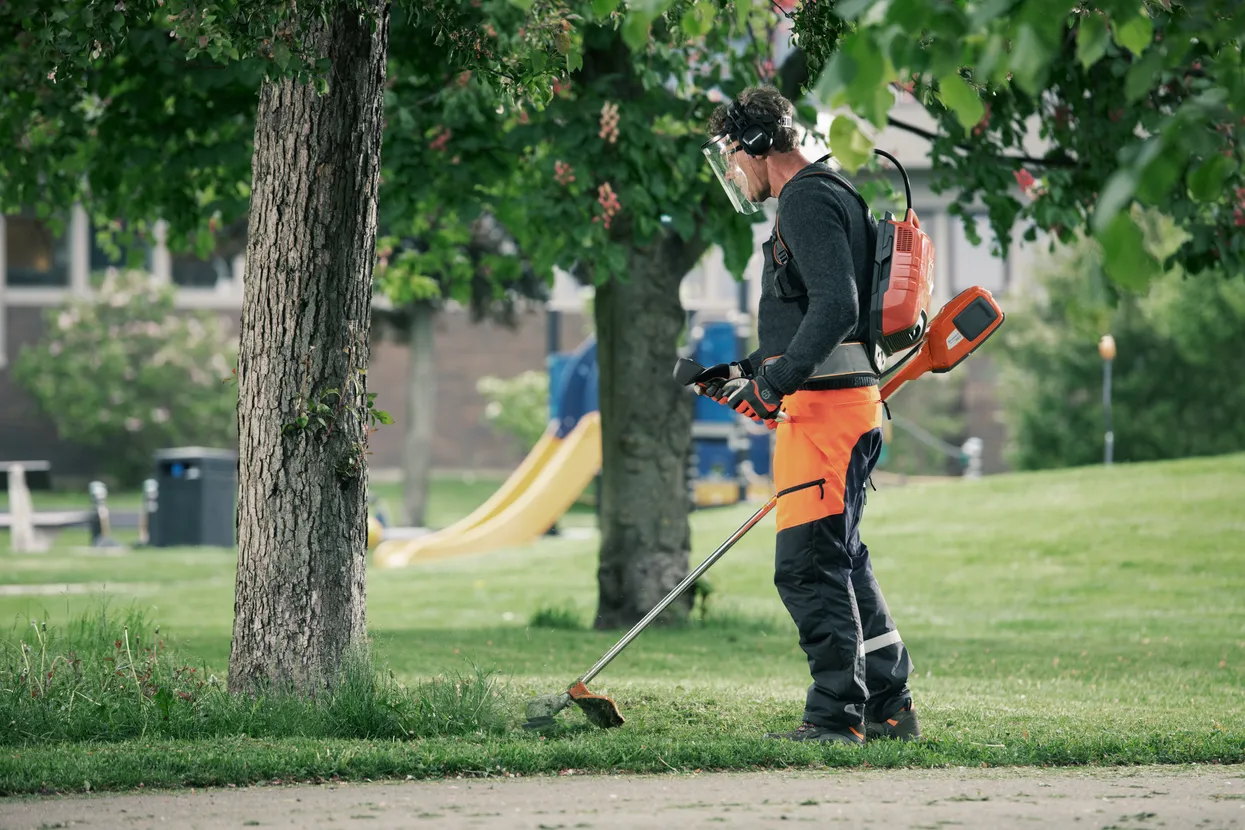 O persoană care folosește o mașină de tuns iarba într-un parc, purtând echipament de protecție de lucru.