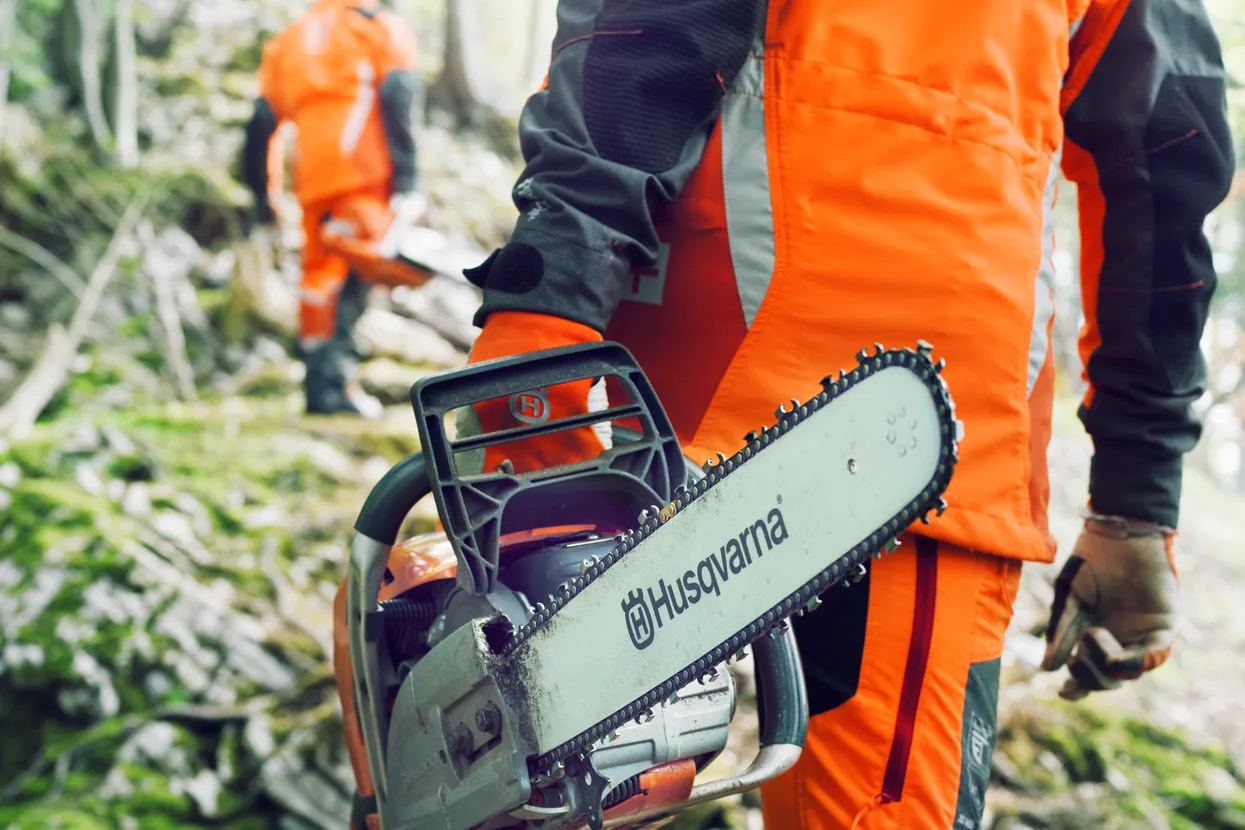 O persoană care ține o drujbă Husqvarna mergând într-o pădure, urmată de o altă persoană.