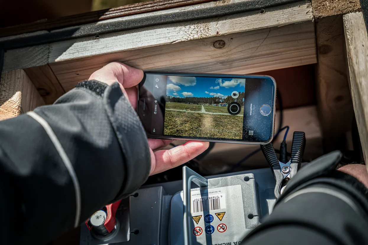 O persoană care face o fotografie a unui gazon cu un smartphone, lângă o baterie cu cleme atașate.