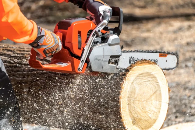 O persoană care folosește un ferăstrău cu lanț Husqvarna pentru a tăia un buștean, cu așchii de lemn zburând.