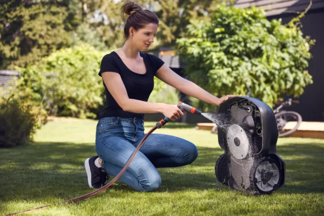 O persoană care curăță o mașină de tuns iarba robotizată cu un furtun de grădină într-o curte.